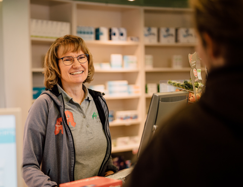 Mitarbeiterin der Medardus Apotheke bedient einen Kunden an der Kasse, im Hintergrund Regale mit Gesundheitsprodukten.